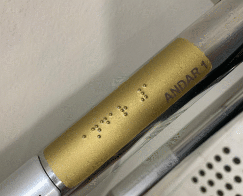 Placa slim de corrimão acessível dourada, com texto em preto e Braille indicando “Andar 1”, instalada sobre corrimão metálico conforme a norma NBR 9050.