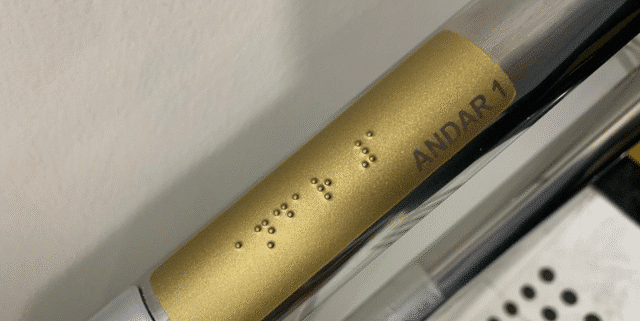 Placa slim de corrimão acessível dourada, com texto em preto e Braille indicando “Andar 1”, instalada sobre corrimão metálico conforme a norma NBR 9050.